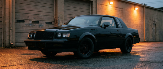 Blacked-out 1987 Buick GNX parked on wet asphalt outside a closed industrial garage