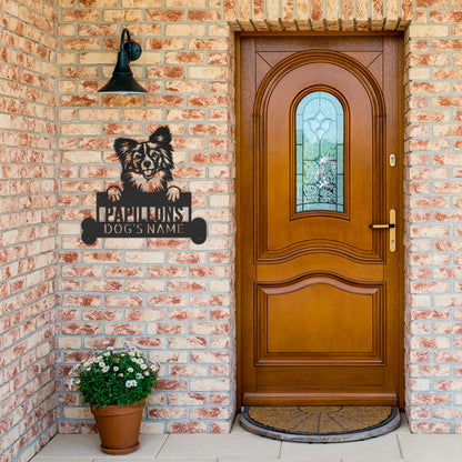 A brick wall with a wooden door, a bell light above, a Custom Papillon Dog Name Sign, Personalized Steel Monogram Wall Art, Pet Home Decor, Animal Lovers Gift, and a flower pot with white flowers on the left.