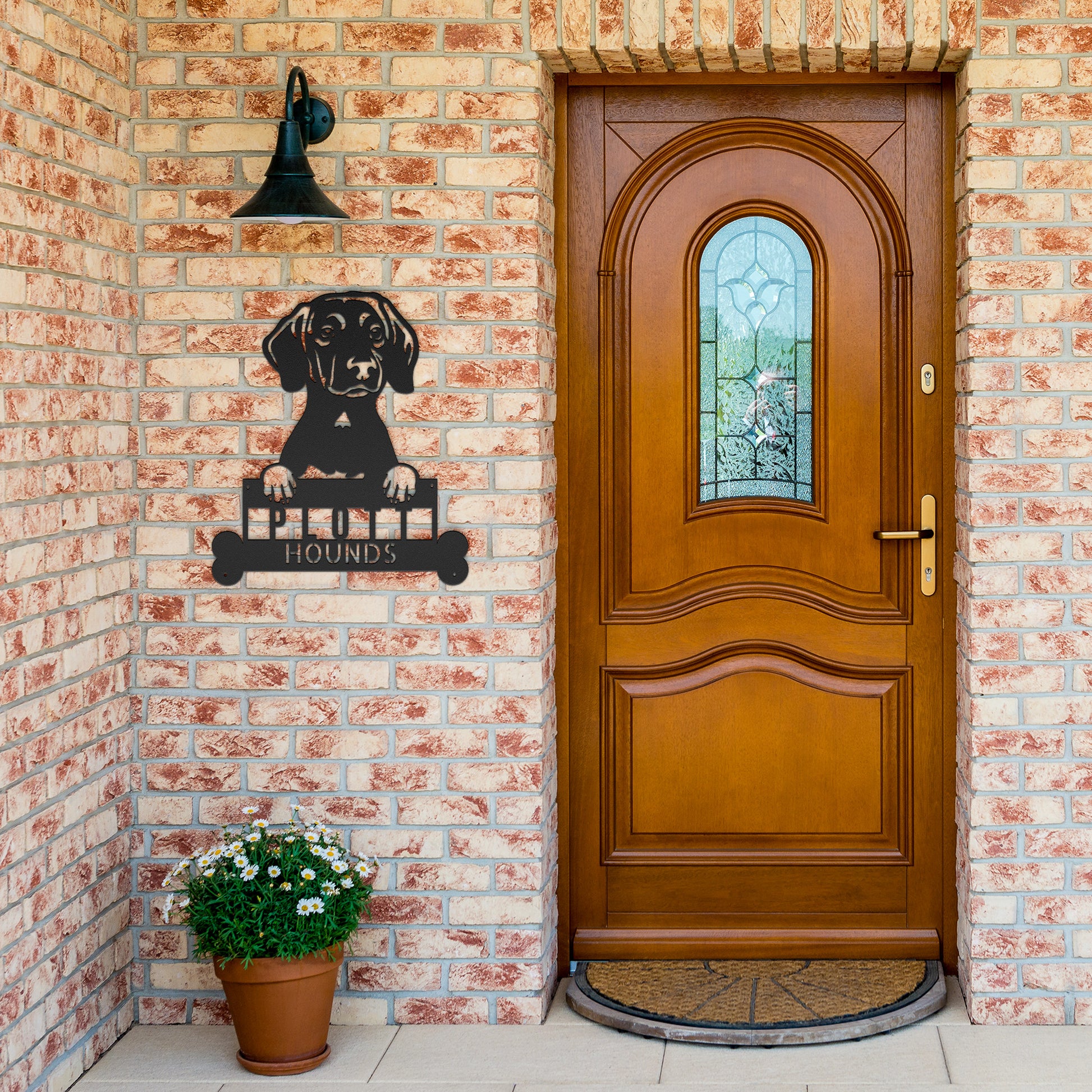 A brick house entrance with a wooden front door, a potted plant with white flowers, and a PITBULL METAL SIGN ARTWORK, Custom Plott Hound Home Décor, Personalized Dog Name Sign, Gift for Animal Enthusiasts featuring a dog's head and the word "HOUNDS" below it.