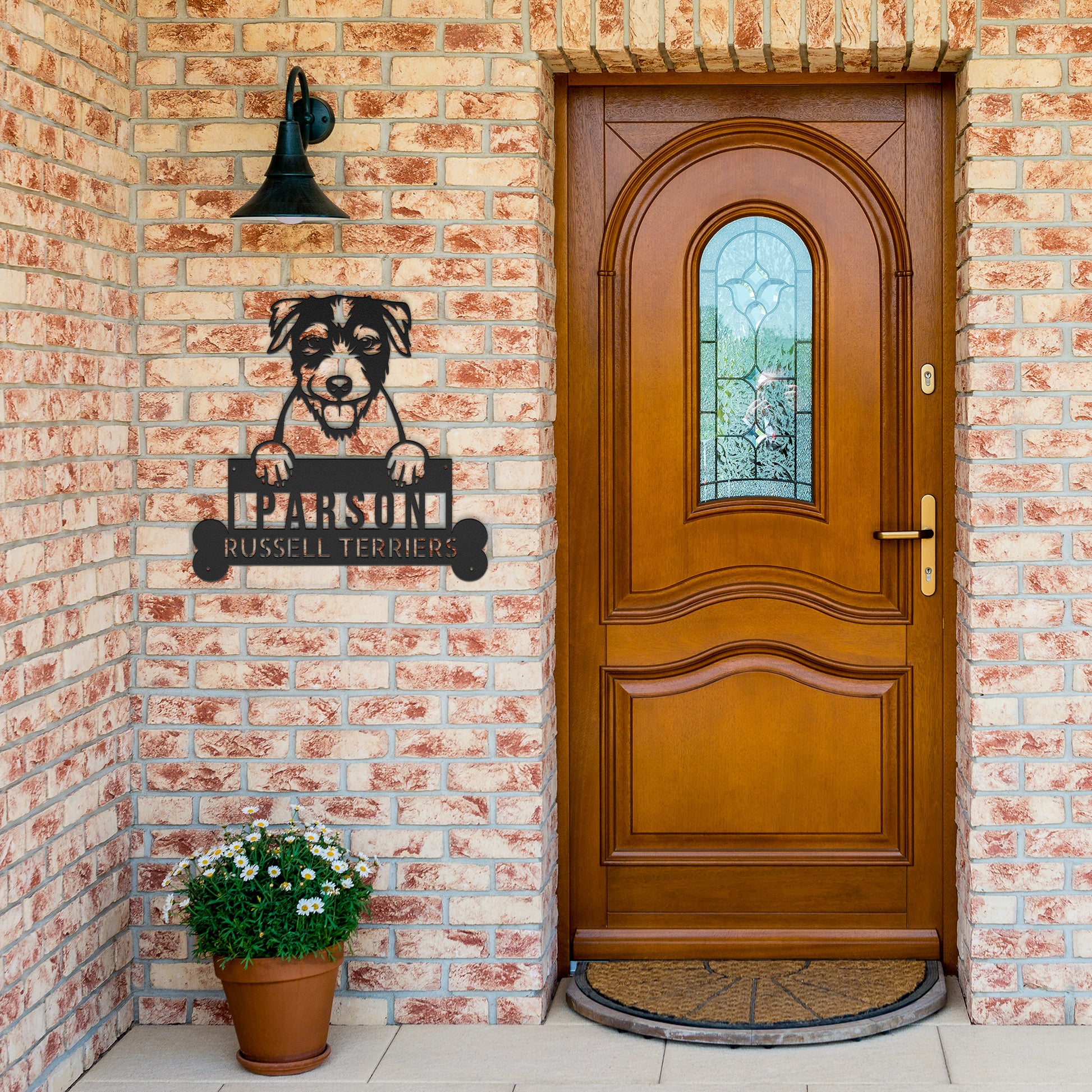 A wooden door with an arched window is set in a brick wall. A potted plant sits on the left, and a Parson Russell Terrier Wall Art, Custom Dog Name Steel Monogram, Personalized Home Decor for Animal Lovers Gift hangs beside the door.