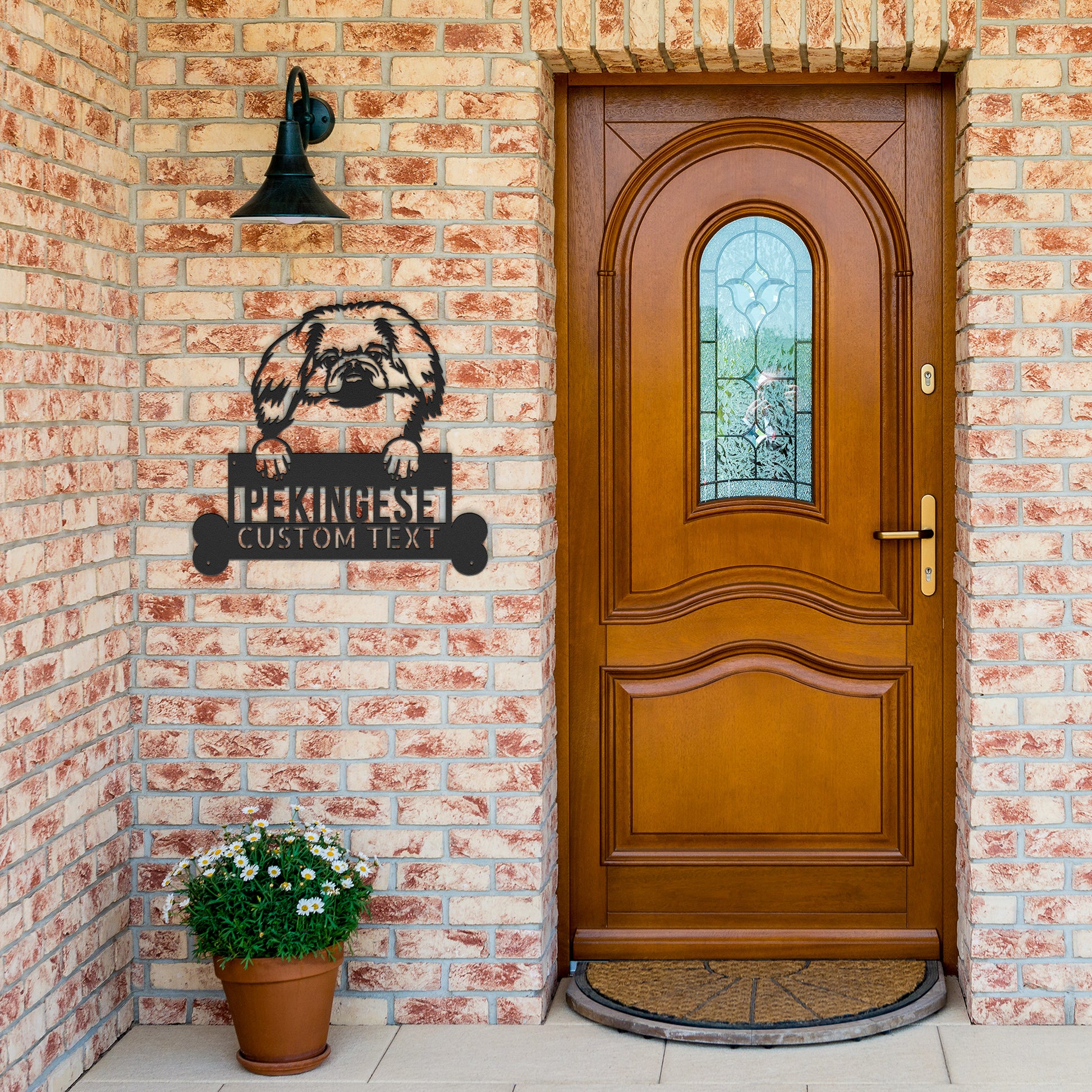 A wooden door with a circular glass insert is set in a brick wall. A Pekingese Dog Name Steel Wall Art, Personalized Monogram Sign for Home Decor, Perfect Animal Lovers Gift is mounted above a potted plant on the doorstep, perfect for those who appreciate unique pet wall art.