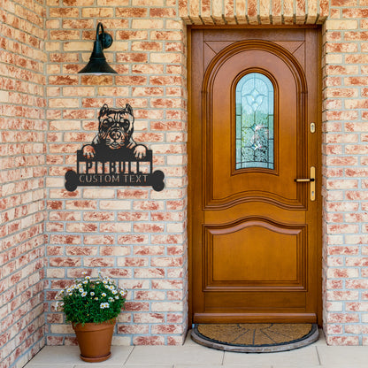 A brick wall front entrance with a wooden door and a doormat. A Pitbull Metal Sign, Personalized Dog Wall Art, Custom Pitbull Name Sign for Home Decor, Perfect Gift for Dog Owners featuring a dog and text is on the wall next to a potted plant with white flowers. The custom dog monogram adds a unique touch to the charming setup.