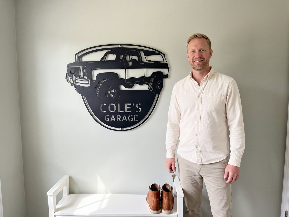 Personalized 1973 K5 Blazer metal sign reading COLE'S GARAGE, hung above a white mudroom bench with work boots as a smiling man holds his keys