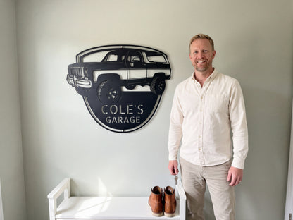 Personalized 1973 K5 Blazer metal sign reading COLE'S GARAGE, hung above a white mudroom bench with work boots as a smiling man holds his keys