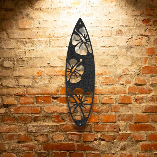 The "Surf Board Flowers - Steel Sign," a black surfboard-shaped metal decoration featuring laser-cut hibiscus flower designs, hangs elegantly on a brick wall with overhead lighting. This powder-coated piece adds sophistication to any space.